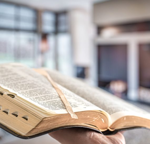 a-man-holds-a-bible-against-the-background-of-the-2021-08-30-09-15-18-utc A man holds a Bible against the background of the living room. Reading a book in a cozy atmosphere. Close up.
